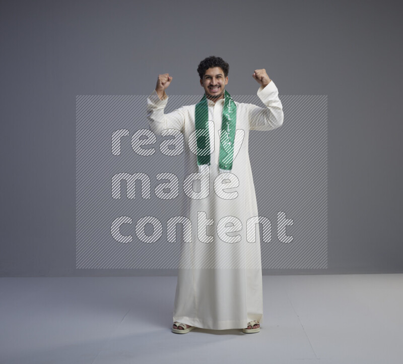 A Saudi man standing wearing thob and Saudi flag scarf on gray background