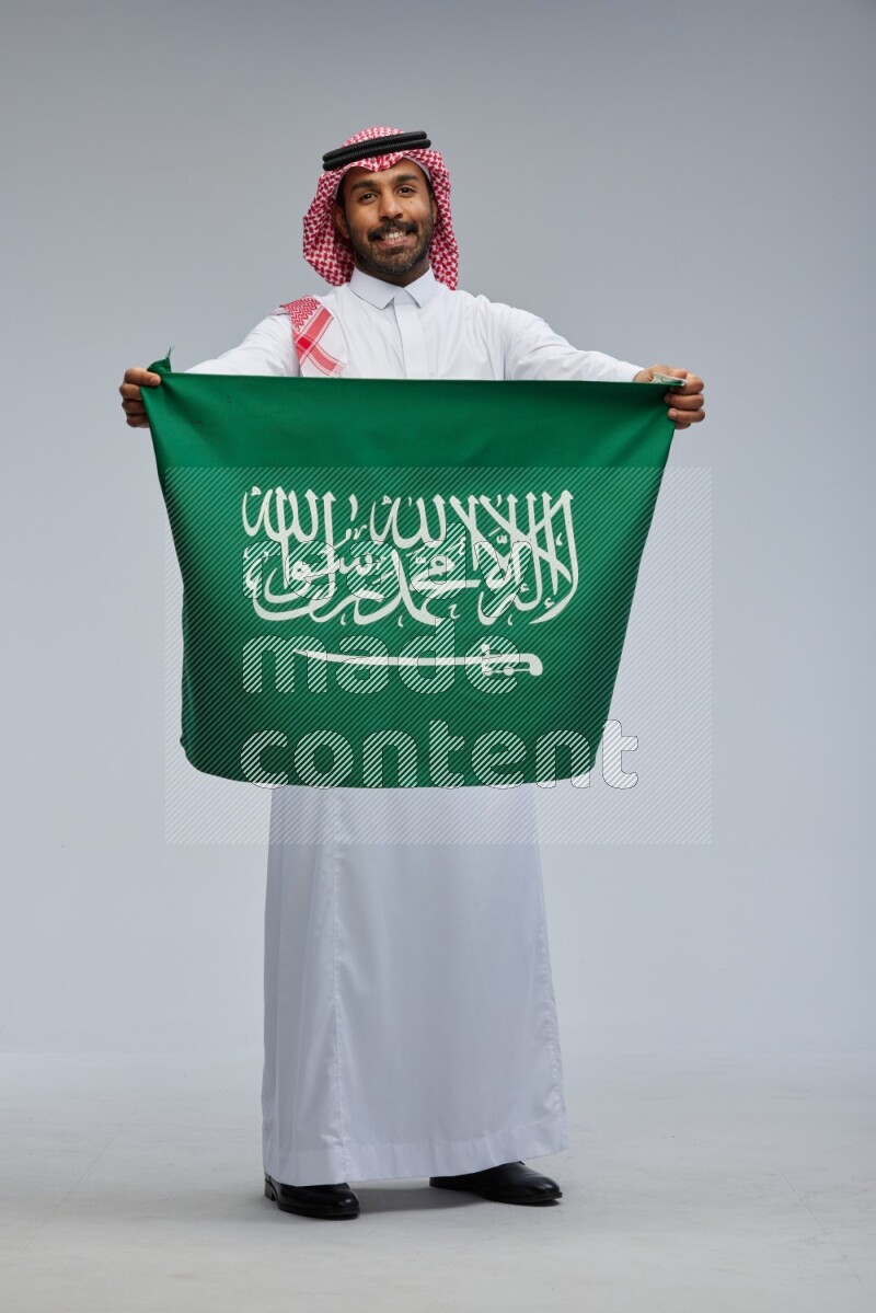 Saudi man Wearing Thob and shomag standing holding Saudi flag on Gray background