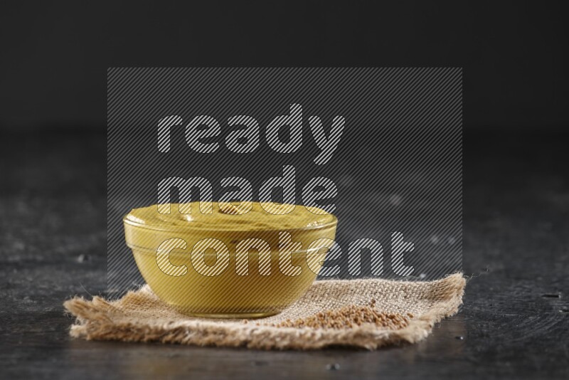 A glass bowl full of mustard paste set on a burlap piece with some mustard seeds on a textured black flooring