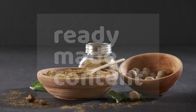 A wooden bowl with a spoon in it and glass spice jar all, full of nutmeg powder and a wooden bowl full of whole nutmeg seeds on a black flooring