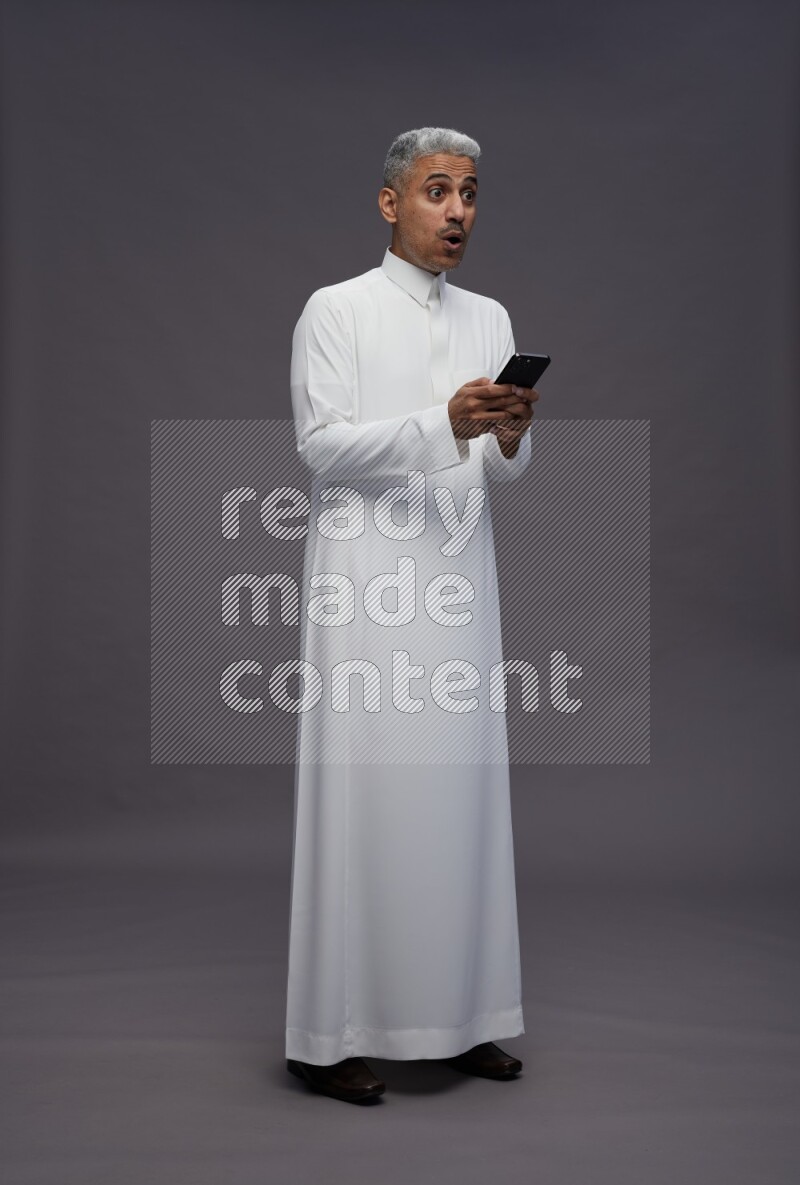 Saudi man wearing thob standing texting on phone on gray background