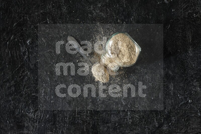 وعاء زجاجي للتوابل مقلوب وملعقة معدنية ممتلئان ببودرة الهيل والبودرة تناثرت منه على خلفية سوداء