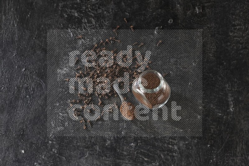 وعاء زجاجي للبهارات وملعقة معدنية ممتلئان ببودرة القرنفل مع حبوب القرنفل المتناثرة على أرضية سوداء