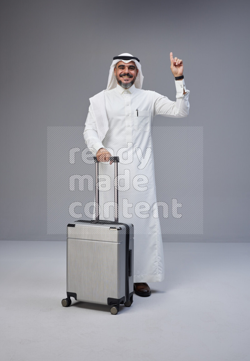 Saudi man wearing Thob and white Shomag standing holding Travel bag on Gray background
