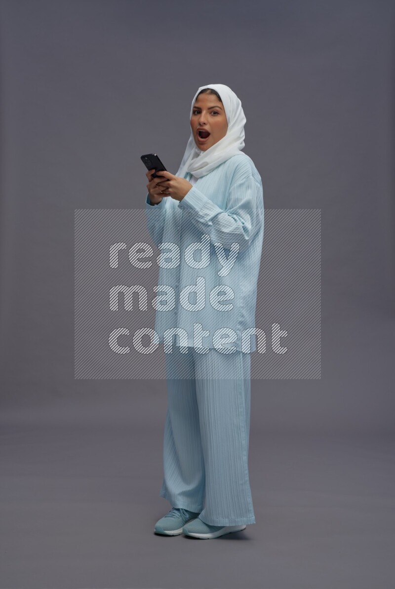 Saudi woman wearing hijab clothes standing texting on phone on gray background