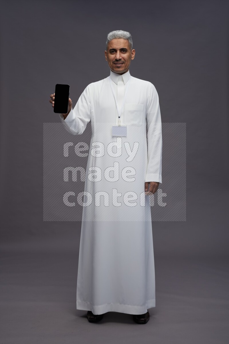 Saudi man wearing thob with neck strap employee badge standing showing phone to camera on gray background