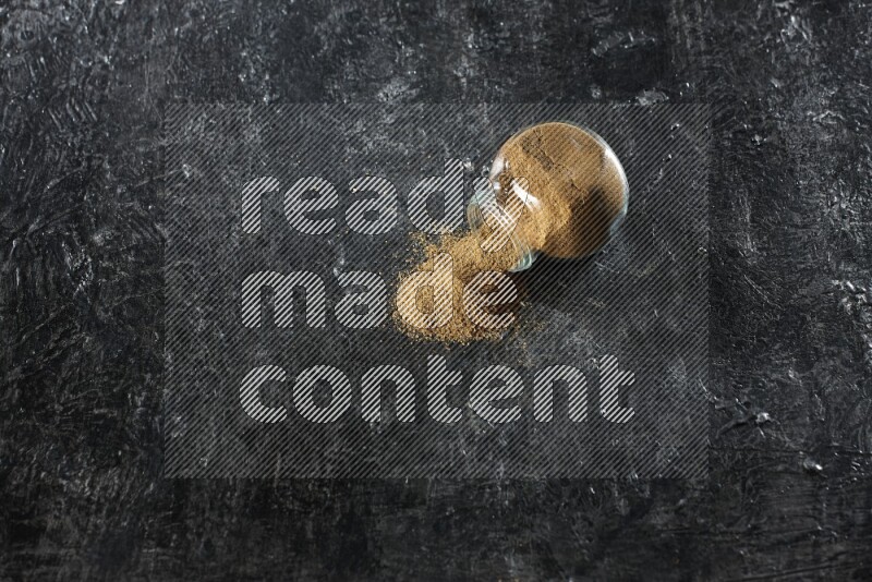 A flipped glass spice jar full of cumin powder and powder spilled out on a textured black flooring