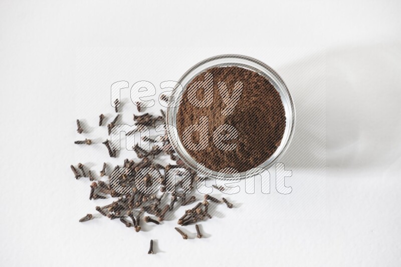 A glass bowl full of cloves powder and cloves grains spread on white flooring
