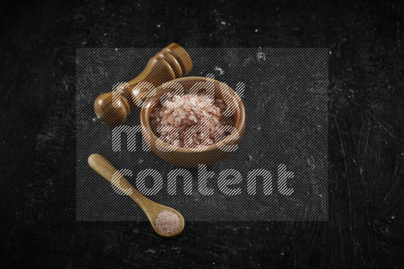 A wooden bowl and spoon filled with coarse pink himalayan salt and a wooden grinder beside them on black background