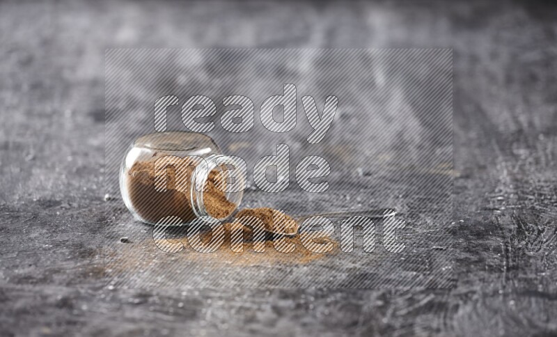 Herbal glass jar full of cinnamon powder flipped and a metal spoon on textured black background