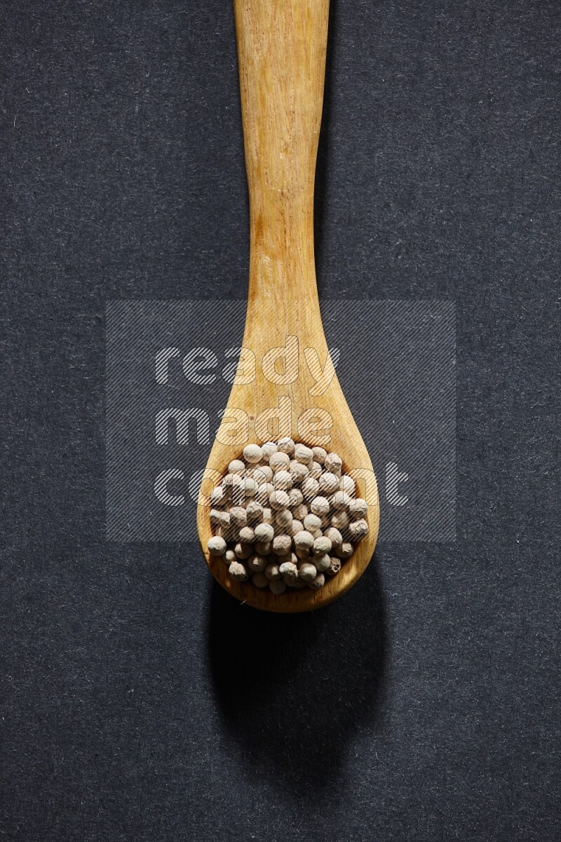 A wooden spoon full of white pepper beads on black flooring