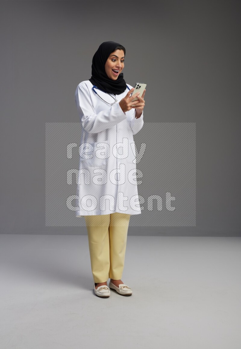 Saudi woman wearing lab coat with stethoscope standing texting on phone on Gray background