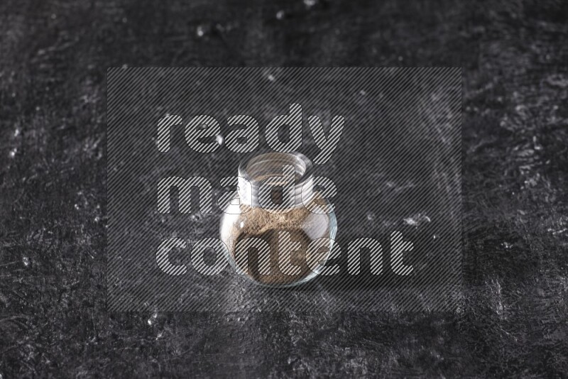 A glass spice jar full of black pepper powder on textured black flooring