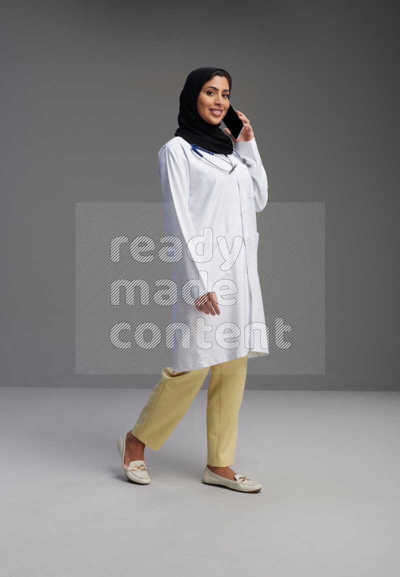 Saudi woman wearing lab coat with stethoscope standing talking on phone on Gray background