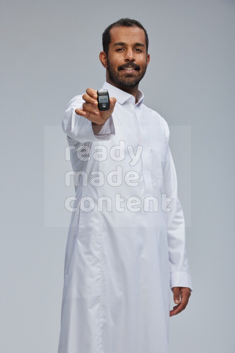 Saudi man wearing thob and shomag standing holding car key on gray background