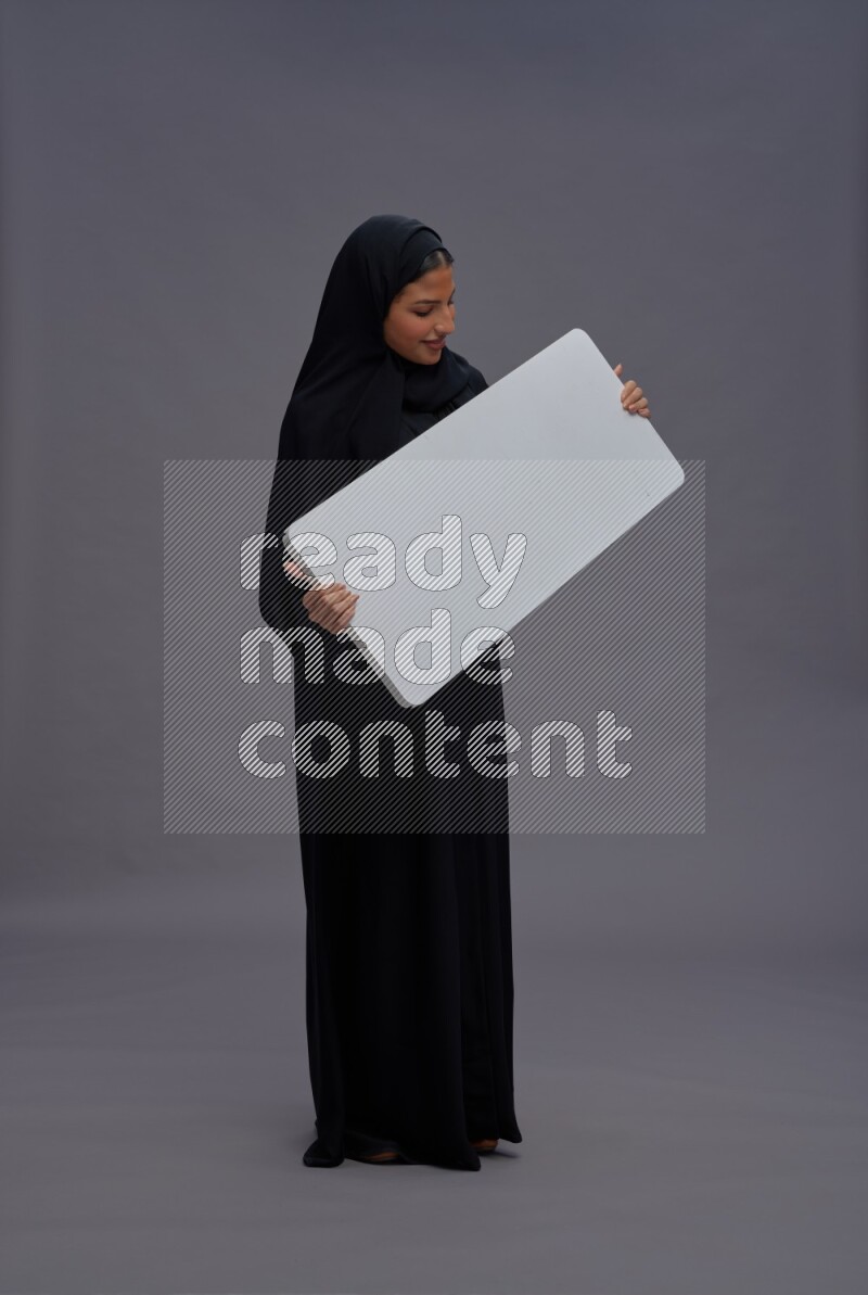Saudi woman wearing Abaya standing holding white board on gray background