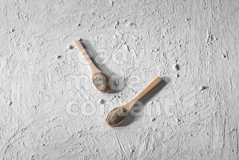 2 wooden spoons full of black pepper powder on a textured white background