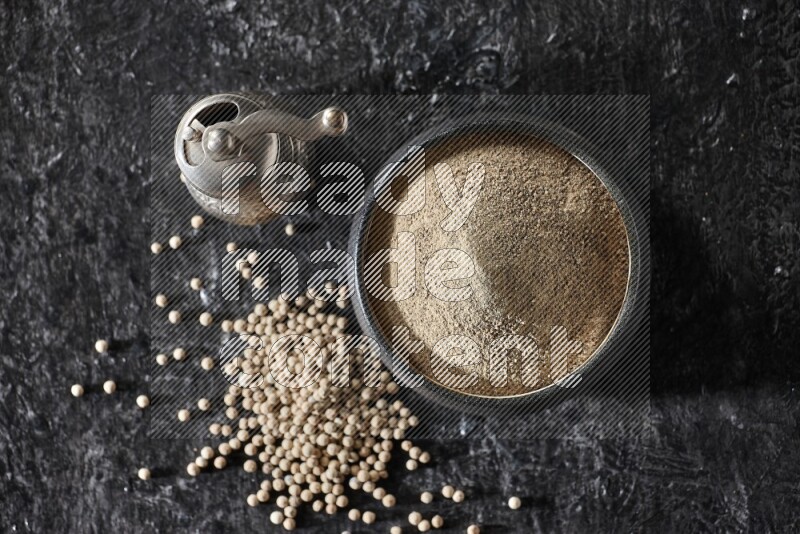 Black pottery bowl full of white pepper powder with pepper beads and metal pepper grinder on textured black flooring