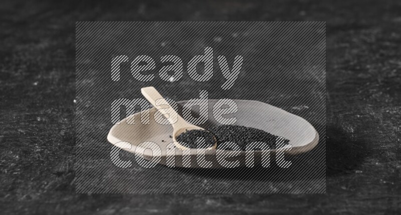 صحن فخاري متعدد الألوان ممتلئ ببذور حبة البركة وملعقة خشبية ممتلئة بالبذور على أرضية سوداء