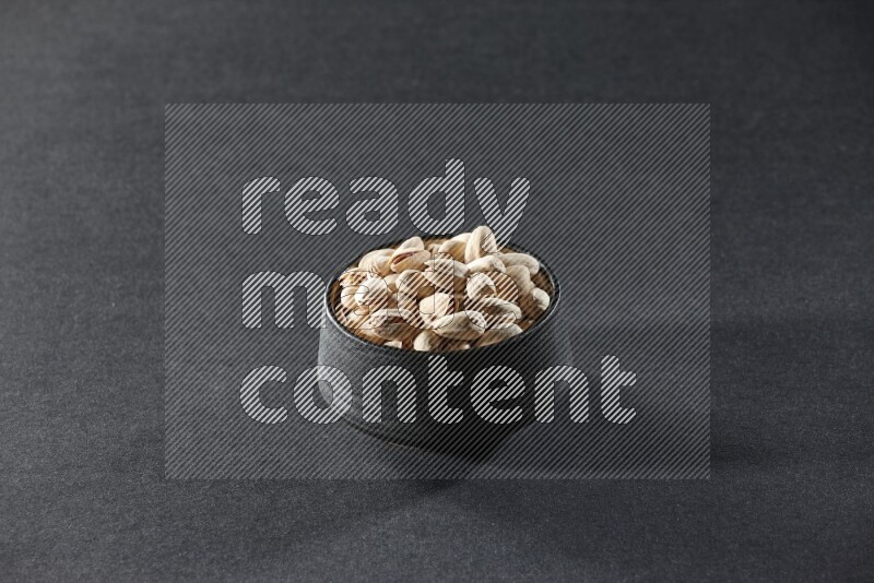 A black pottery bowl full of pistachios on a black background in different angles