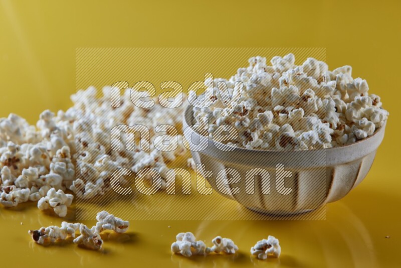 A white pottery bowl full of popcorn with popcorn beside it on a yellow background in different angles