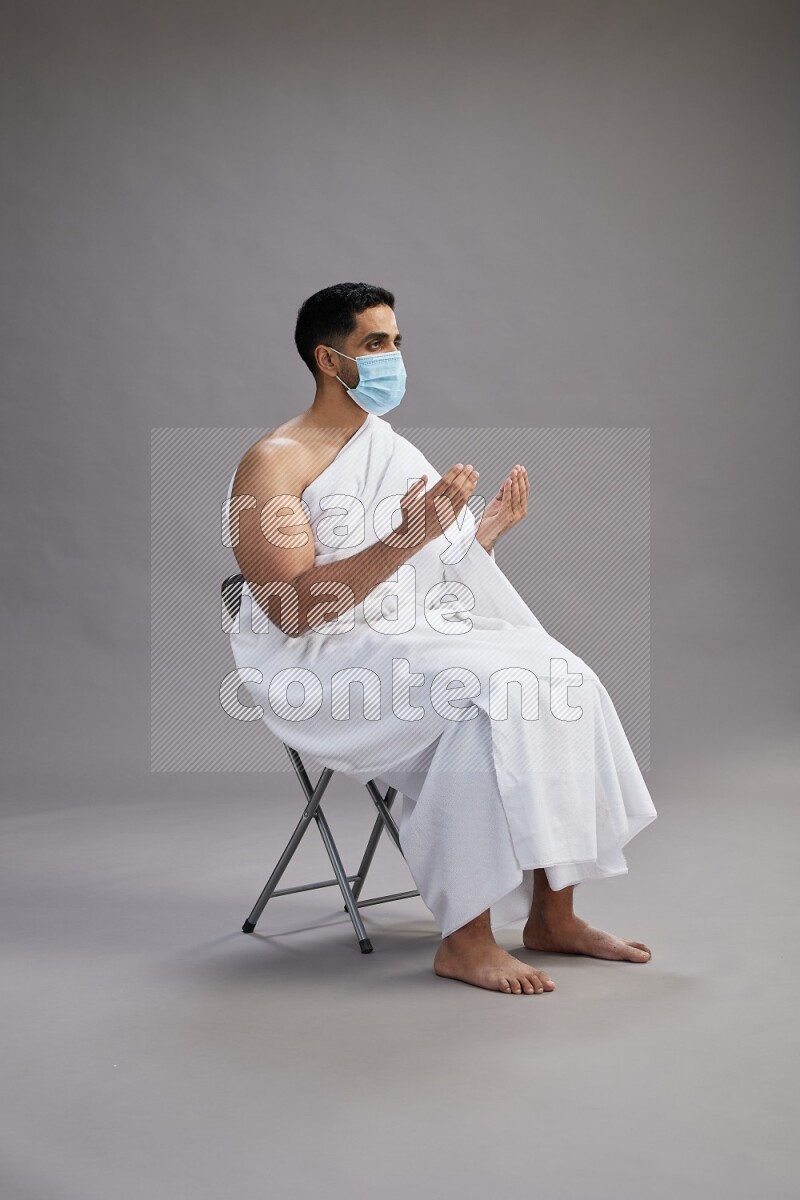 A man wearing Ehram with face mask sitting on chair dua'a on gray background