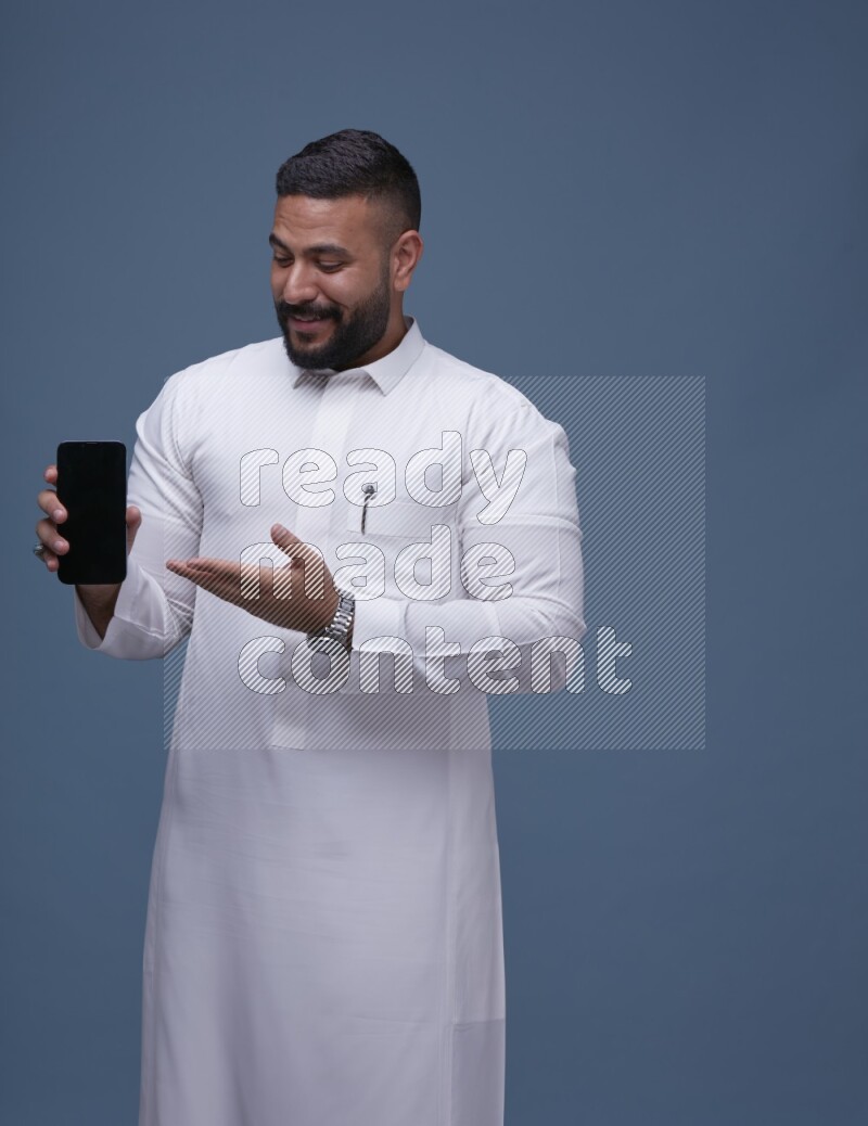 A man Showing his phone in Blue Background wearing Saudi Thob